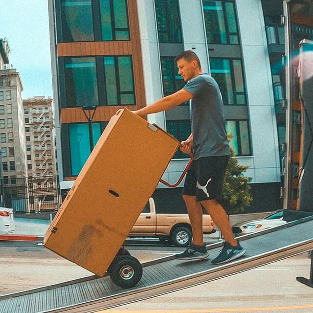 Next Moving professional apartment movers are walking down the ramp out of the moving trucks, holding hand truck dolly and with wardrobe boxes full of moving blankets on it . The box truck is parked at the front of the apartment complexes.