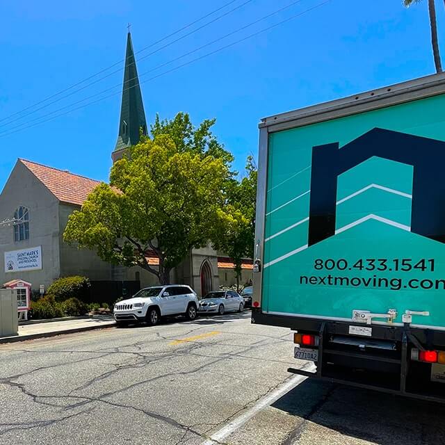Next Moving 20-foot box truck with branded decals on it and local Glendale movers is parked by an apartment complex across the street from St Mark's Episcopal Church in Glendale, California.