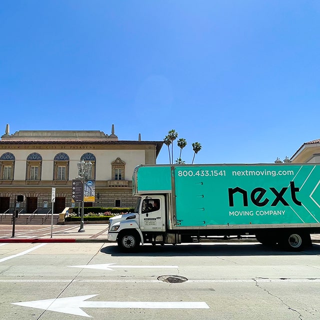 Next Moving local movers inside 28-foot box truck with attic with Next Moving corporative decals on it passing by The Pasadena Civic Auditorium on their way to client's delivery address. The decal on the side of the truck shows 800.433.1541 phone number nextmoving.com website and large Next Moving logo on branded mint color background.
