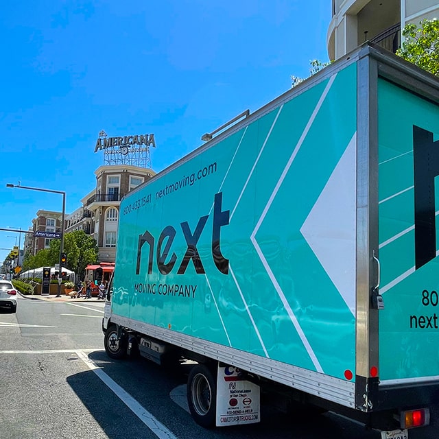 Next Moving 20-foot box truck with branded decals on it and long-distance movers inside is waiting for a green light at the intersection across the street from the Americana at the Brand in Glendale CA.