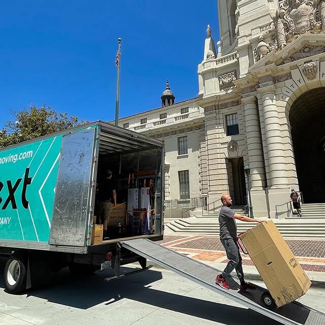 Next Moving 28-foot box truck with attic and Next Moving corporative decals on it is parked at the front of the Pasadena City Hall with opened back doors and the walking ramp pooled out. The decal on the side of the truck shows 800.433.1541 phone number nextmoving.com website and a large Next Moving logo on branded mint color background. One professional Pasadena local mover is pooling a hand truck with a wardrobe box on into the truck. Another mover is stacking items inside the truck for safe transportation.