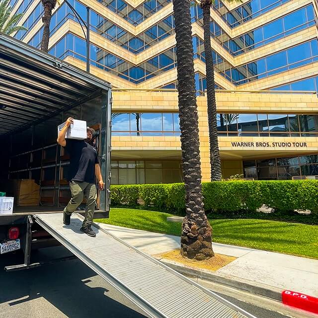 Next Moving commerical and office mover walking down the ramp from the moving truck with the box on his shoulder in the middle of the unloading process at the Warner Bros. Studio Tour Hollywood Building located in Burbank California.