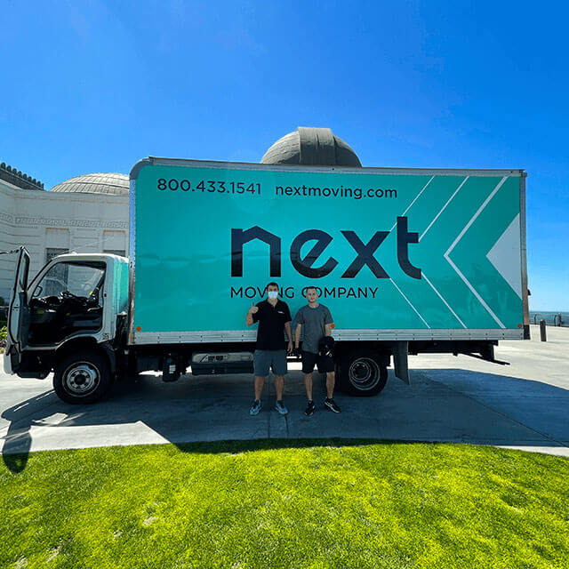 Two professional local movers standing at the front of the middle of the Next Moving box truck during the moving process. The truck is parked at the Griffith Observatory, Los Angeles, California. The body of the truck has Next Moving Company logo on it with the phone number 8004331541, and email address nextmoving.com.
